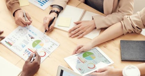 People working at a table pointing with pens at a document laying in the center of the table group. Also, a digital version of the document is displayed on an Ipad in one of the group member's hands. 