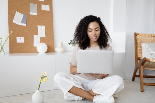 A young woman sits cross-legged in a bright, modern room, smiling while working on her laptop. A corkboard with notes and a vase with flowers are in the background.