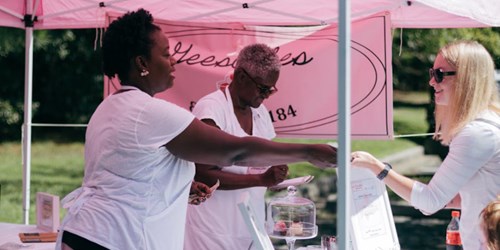 A vendor at an outdoor market hands a bag to a customer under a pink tent, with another vendor arranging cupcakes nearby.