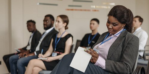 A woman in a business suit takes notes during a professional conference, with other attendees listening in the background.