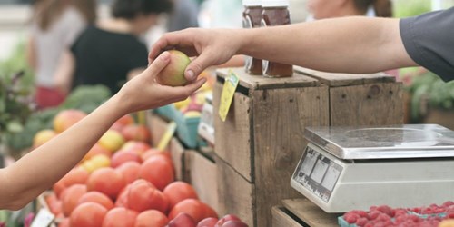A vendor hands a fresh apple to a customer at an outdoor farmers' market, with colorful produce in the background.